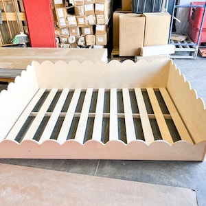 May include: A light-colored wooden bed frame with a scalloped edge design. The bed frame has a slatted base and is made of unfinished wood. The frame is rectangular and sits on a concrete floor.