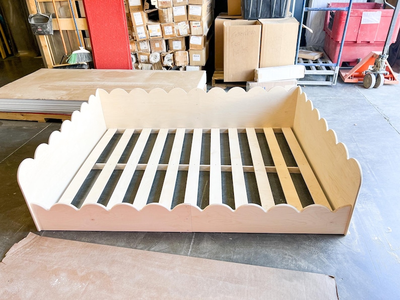 May include: A light-colored wooden bed frame with a scalloped edge design. The bed frame has a slatted base and is made of unfinished wood. The frame is rectangular and sits on a concrete floor.
