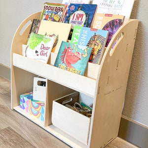 May include: A light-colored wooden bookshelf with a curved design, holding various children's books. The books have colorful covers, including titles like "Snow Day," "I Spy Spooky Night," and "Dear Girl." The bookshelf also has storage compartments for additional items.