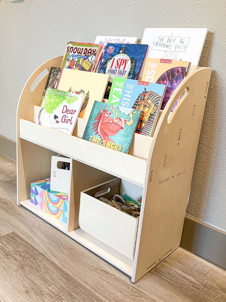 May include: A light-colored wooden bookshelf with a curved design, holding various children's books. The books have colorful covers, including titles like "Snow Day," "I Spy Spooky Night," and "Dear Girl." The bookshelf also has storage compartments for additional items.