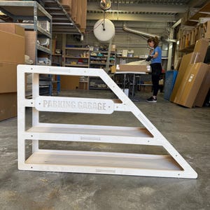 May include: A wooden toy parking garage with three shelves and a triangular side. The text "PARKING GARAGE" is displayed on the front. The structure is made of light-colored wood and is in an industrial setting.