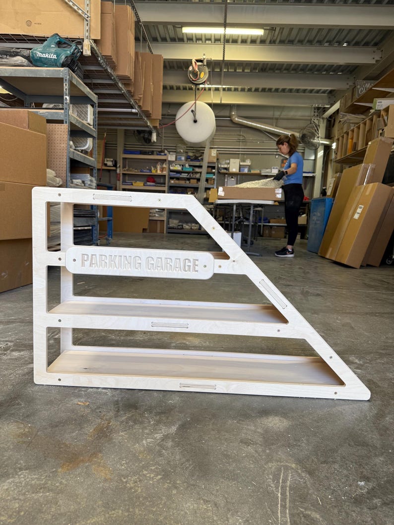 May include: A wooden toy parking garage with three shelves and a triangular side. The text "PARKING GARAGE" is displayed on the front. The structure is made of light-colored wood and is in an industrial setting.
