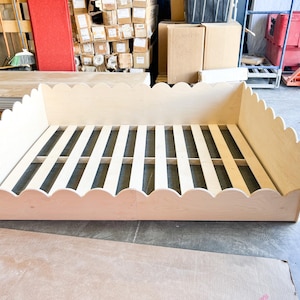 May include: A light-colored wooden bed frame with a scalloped edge design. The bed frame has a slatted base and is made of unfinished wood. The frame is rectangular and sits on a concrete floor.