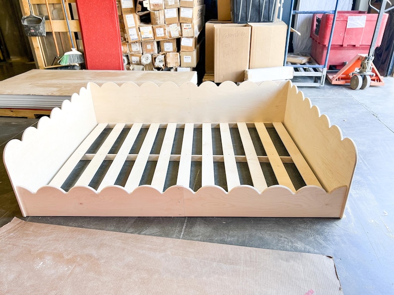 May include: A light-colored wooden bed frame with a scalloped edge design. The bed frame has a slatted base and is made of unfinished wood. The frame is rectangular and sits on a concrete floor.