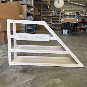 May include: A wooden parking garage shelf unit with three levels. The unit is light-colored and features the words "PARKING GARAGE" in a horizontal sign. The unit has an angled design and is made of wood.