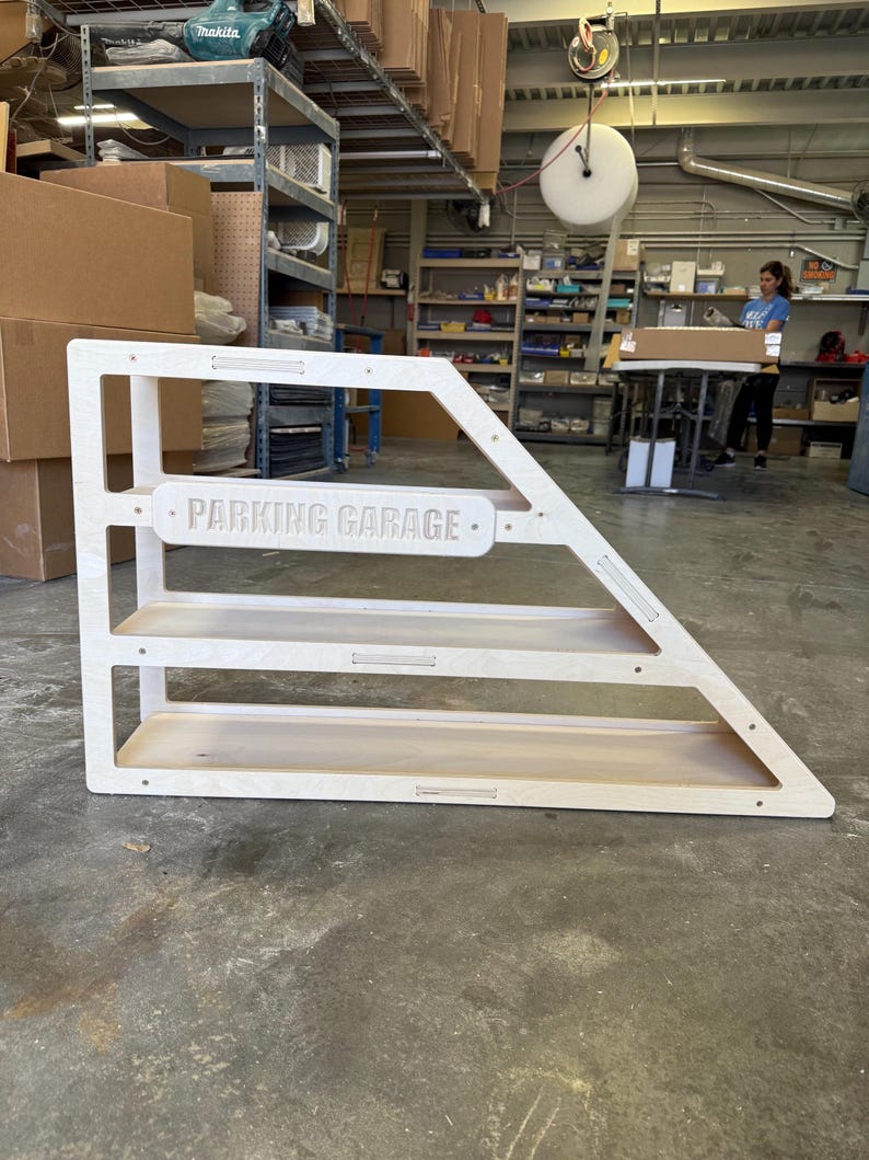 May include: A wooden parking garage shelf unit with three levels. The unit is light-colored and features the words "PARKING GARAGE" in a horizontal sign. The unit has an angled design and is made of wood.