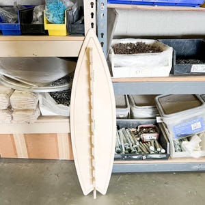 May include: A light wood surfboard-shaped organizer with slots for holding various items. The organizer is standing upright on a concrete floor in front of a metal shelving unit.