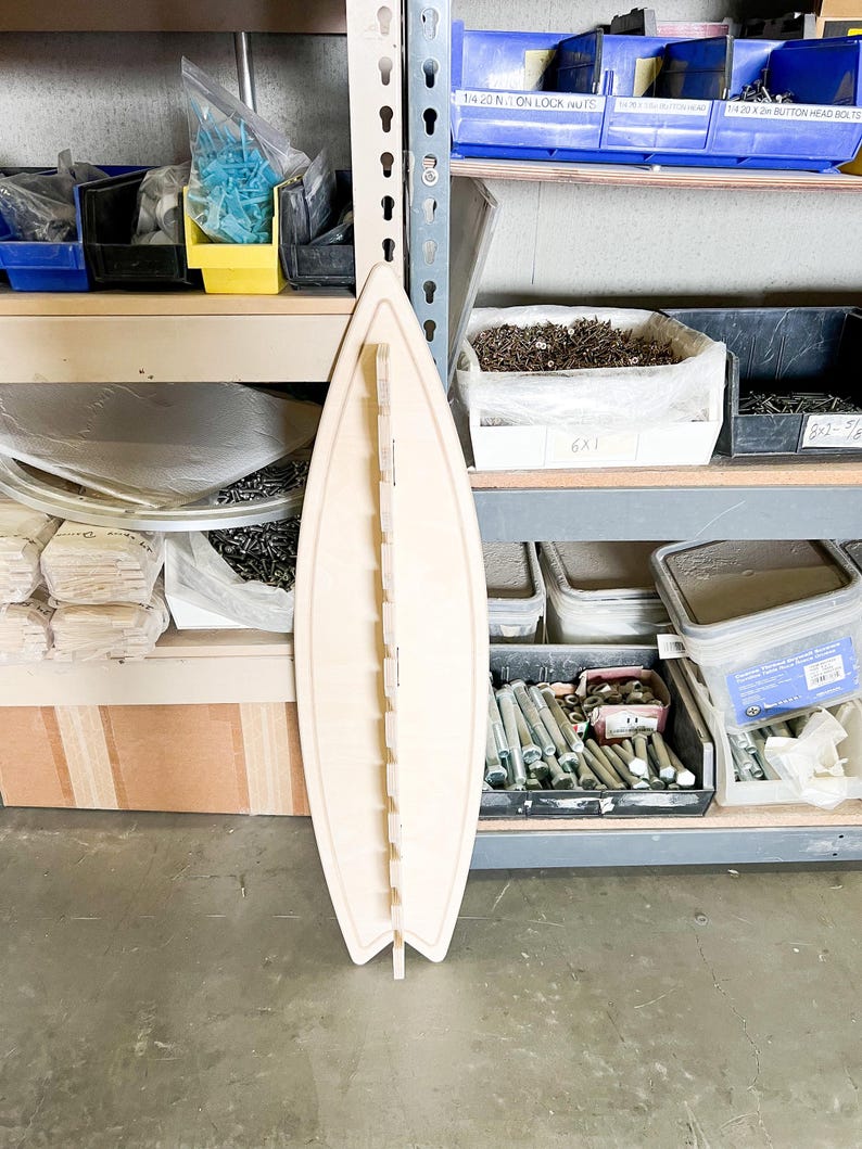May include: A light wood surfboard-shaped organizer with slots for holding various items. The organizer is standing upright on a concrete floor in front of a metal shelving unit.