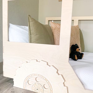 May include: A white wooden toddler bed with a gear-shaped design on the side. The bed has a white mattress and a green and brown pillow on it. A black teddy bear is sitting on the bed.