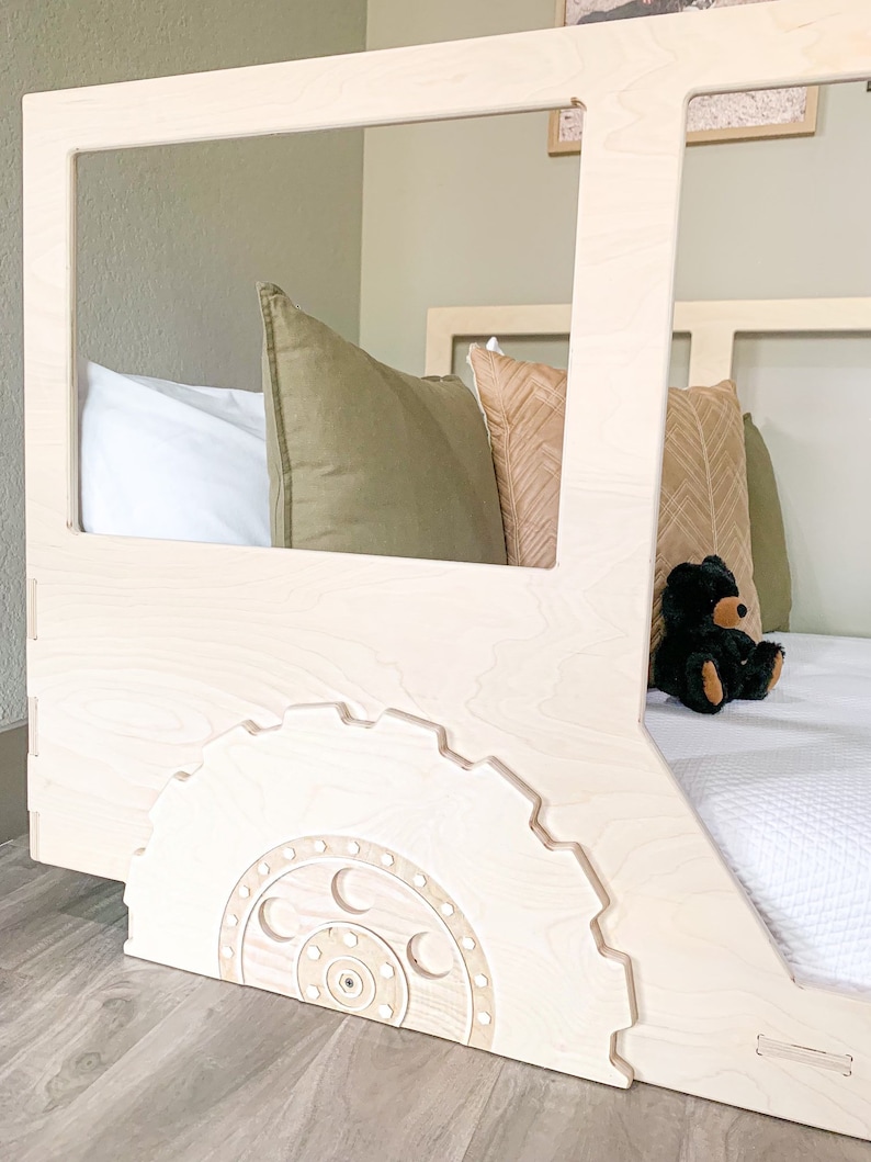 May include: A white wooden toddler bed with a gear-shaped design on the side. The bed has a white mattress and a green and brown pillow on it. A black teddy bear is sitting on the bed.