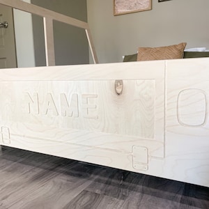 May include: A white wooden bed frame with a cutout of the word "NAME" on the headboard. The bed frame has a simple design with rounded edges and a light wood finish.