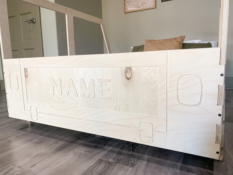 May include: A white wooden bed frame with a cutout of the word "NAME" on the headboard. The bed frame has a simple design with rounded edges and a light wood finish.