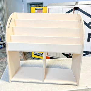 May include: A light-colored wooden bookshelf with multiple shelves and rounded handles. The bookshelf has a rectangular base with two compartments and several horizontal shelves for storage. The wood has a natural, unfinished appearance.