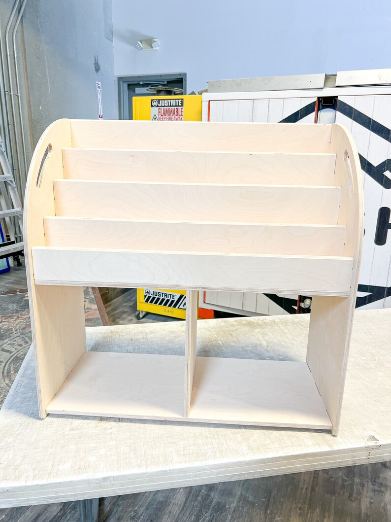 May include: A light-colored wooden bookshelf with multiple shelves and rounded handles. The bookshelf has a rectangular base with two compartments and several horizontal shelves for storage. The wood has a natural, unfinished appearance.