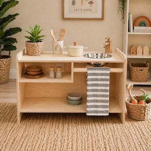 May include: A wooden play kitchen with a stainless steel sink, a small pot, and a striped towel. It includes wooden utensils, plates, bowls, and salt and pepper shakers. A framed print that says "LET'S COOK" hangs above the kitchen.
