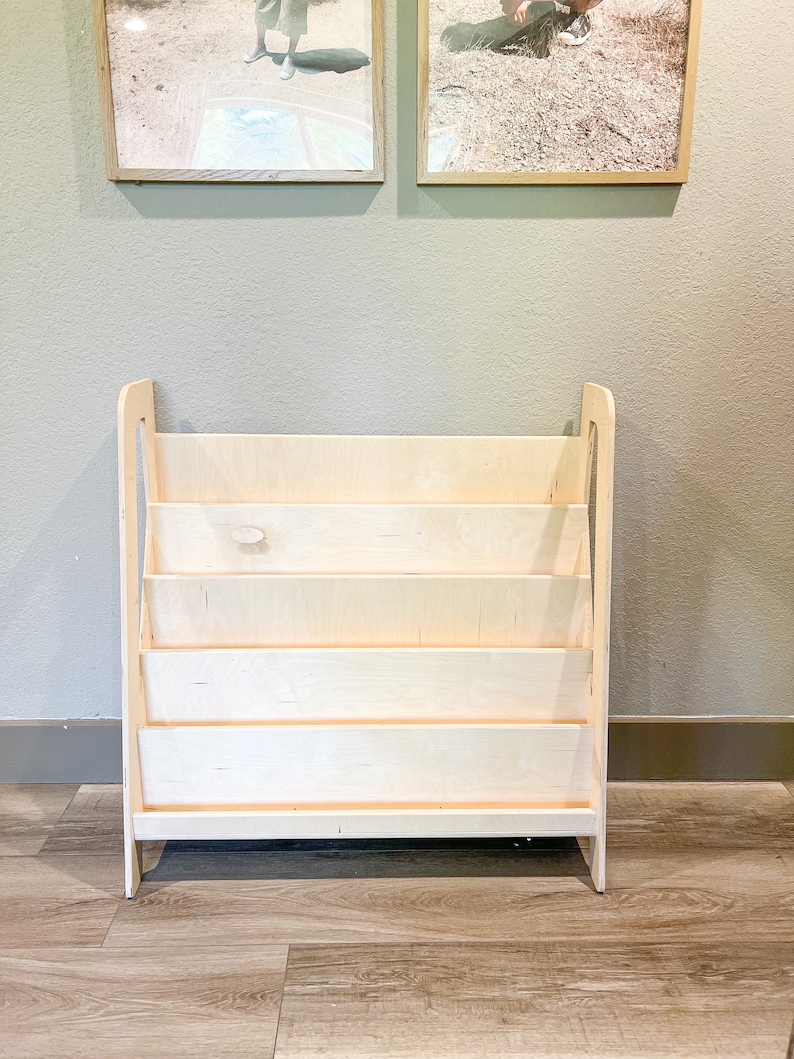May include: A light-colored wooden bookshelf with four shelves. The bookshelf has a simple, modern design with a rectangular frame and a slatted back. The shelves are evenly spaced and appear to be made of the same light-colored wood. The bookshelf is standing on a wooden floor.