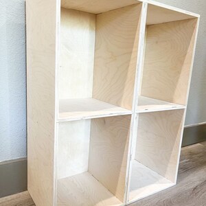 May include: A wooden shelf unit with four open compartments. The shelf is made of light-coloured wood and has a simple, modern design.
