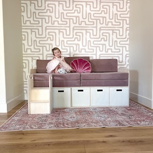 May include: A pink and white upholstered daybed with storage compartments and a built-in step. The daybed is against a wall with a pink and white geometric wallpaper. A young girl is sitting on the daybed.