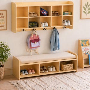 May include: A light wood storage bench with a cushioned top, paired with a matching wall-mounted shelf unit. The shelf has multiple compartments for shoes and other items. A backpack and coat hang below. A framed sign reads "KIND PEOPLE ARE MY KIND OF PEOPLE."