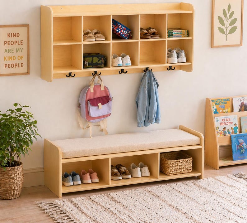 May include: A light wood storage bench with a cushioned top, paired with a matching wall-mounted shelf unit. The shelf has multiple compartments for shoes and other items. A backpack and coat hang below. A framed sign reads "KIND PEOPLE ARE MY KIND OF PEOPLE."