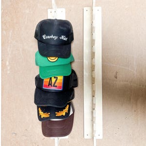 May include: A wooden hat rack with five baseball caps hanging on it. The caps are black, brown, green, black with a rainbow "AZ" logo, and black with the words "Cowboy Hat" in white.