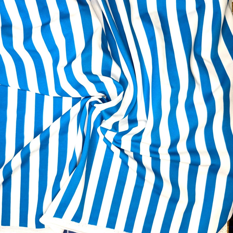 May include: A close-up shot of a fabric with vertical stripes in bright blue and white. The fabric is draped and folded, creating a textured effect. The stripes are evenly spaced and run the length of the material.