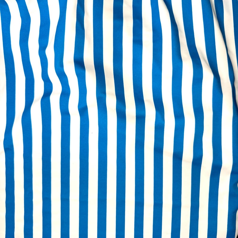 May include: Close-up of a fabric with vertical stripes. The stripes alternate between bright blue and white. The fabric appears to be slightly wrinkled, creating soft curves in the pattern. The overall effect is a clean, nautical aesthetic.