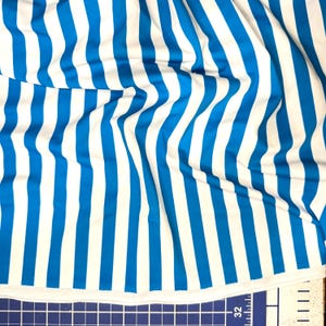 May include: Close-up of a fabric with vertical stripes in bright blue and white. The fabric is slightly wrinkled, showing the texture and flow of the material. The stripes are evenly spaced and run the length of the fabric.
