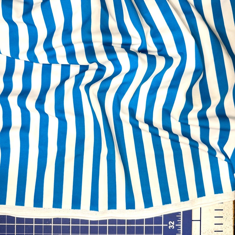 May include: Close-up of a fabric with vertical stripes in bright blue and white. The fabric is slightly wrinkled, showing the texture and flow of the material. The stripes are evenly spaced and run the length of the fabric.