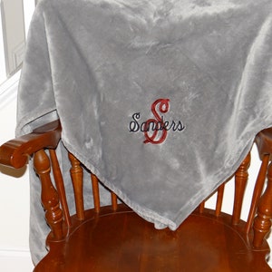 May include: A gray fleece throw blanket with the name "Sanders" embroidered in red and black on the front. The blanket is draped over a wooden chair.