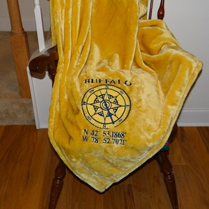 May include: A yellow fleece blanket with blue embroidery. The embroidery includes the word "BUFFALO" and a compass with the coordinates "N 42° 53.1868' W 78° 52.7021'".