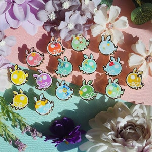 Peut inclure: Une collection de 16 épingles en émail colorées représentant différents personnages de dessins animés. Les épingles sont disposées sur un fond rose clair et bleu avec des fleurs.