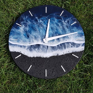 Puede incluir: Un reloj redondo con fondo negro y agujas blancas. La esfera del reloj presenta un diseño artístico de resina azul y blanca de olas del océano rompiendo en una playa de arena.