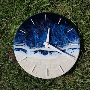 May include: A round clock with a white face and blue and white resin art depicting ocean waves. The clock hands are white.