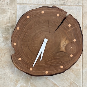 May include: A wooden clock with a white hand and copper-coloured accents. The clock face is a tree trunk slice with visible rings.