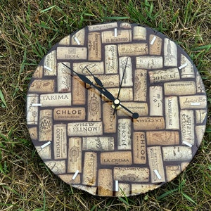 May include: A round clock with a black face and black hands. The clock face is covered in a collage of wine corks with various wine labels printed on them. The labels include names like "Chloe", "Tarina", "Montes Alpha", "La Crema", and "J. Lohr".