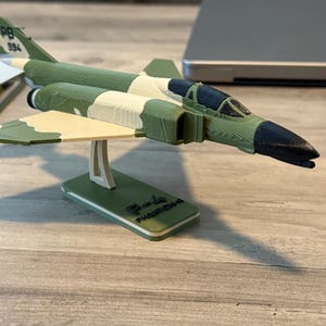 May include: A green, white, and tan model airplane, the F-4 Phantom II, is displayed on a stand. The model has a black nose and cockpit, with the text "PB 994" on the tail. The stand has the text "F-4 Phantom II" on it.