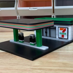 May include: A miniature model of a 7-Eleven convenience store with a green awning and a black base. The store has a white facade with a red and white 7-Eleven logo. There are two gas pumps in front of the store.