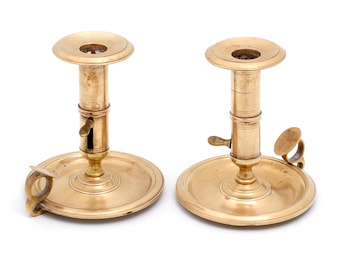 Puede incluir: Dos candelabros antiguos de latón. Cada candelabro tiene una base redonda, una columna central y una copa superior para sostener una vela. Uno tiene un asa y una pequeña tapa con bisagras. El latón tiene un tono dorado cálido.