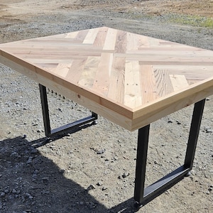 May include: A square wooden table with a light-colored, natural wood surface. The tabletop is constructed from various wood planks arranged in a geometric pattern. The table has black metal legs. The table is approximately 40 inches wide.