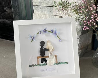May include: A framed pebble art piece featuring a couple on a bench, with a floral arrangement above. The figures are made of black pebbles, the bride's dress is white, and the bench is brown. The frame is white, and the text "Mike & Tricia" is visible.