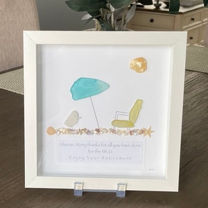 May include: A white framed picture with a sea glass art piece depicting a beach scene with a bird, an umbrella, a chair, and a starfish. The picture includes the text "Sharon, Many thanks for all you have done for the NICU. Enjoy Your Retirement."
