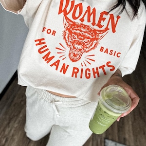 May include: Cream-coloured crop top with the text "WOMEN FOR HUMAN BASIC RIGHTS" in red, featuring a wolf graphic. Paired with light grey sweatpants and a green beverage in a clear plastic cup.