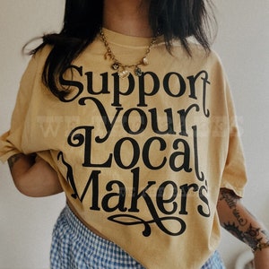 May include: Mustard yellow t-shirt with the text "Support Your Local Makers" in black script. The shirt is paired with blue and white checkered shorts. A gold necklace with charms is visible.
