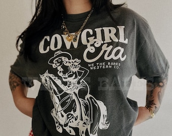 May include: A dark gray t-shirt with a white graphic of a woman riding a horse. The graphic includes the text "Cowgirl Era" and "We the Babes Western Co."