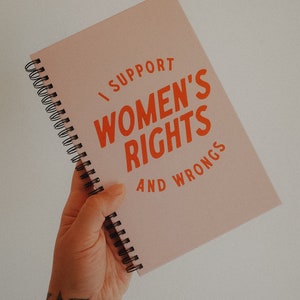 K&ouml;nnte beinhalten: Ein rosa Spiralnotizbuch mit dem Text "I SUPPORT WOMEN'S RIGHTS AND WRONGS" in Orange. Das Notizbuch wird in einer Hand mit einem goldenen Armband und floralen T&auml;towierungen gehalten.