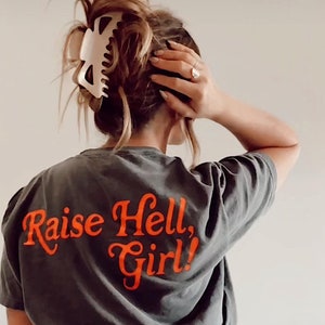 May include: A dark gray t-shirt with the text "Raise Hell, Girl!" printed in orange on the back. The shirt is being worn by a person with their hair pulled back in a bun.