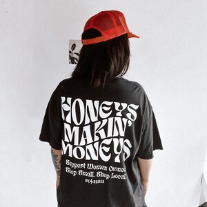 May include: Black t-shirt with white text that reads "Honeys Makin' Moneys" and "Support Women Owned Shop Small Shop Local" with a small lightning bolt and the words "We ⚡️ Babes".