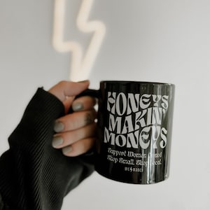 May include: Black ceramic mug with the text "Honeys Makin' Moneys" in white. The mug also has the text "Support Woman Owned. Shop Small. Shop Local." and the logo "We ⚡️ Babes".
