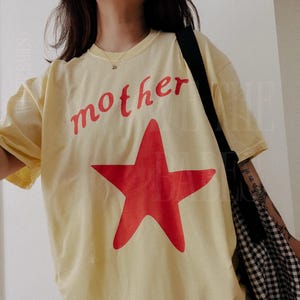 May include: A light yellow t-shirt with the word "mother" in red script above a large red star. The shirt is a casual style, with short sleeves.
